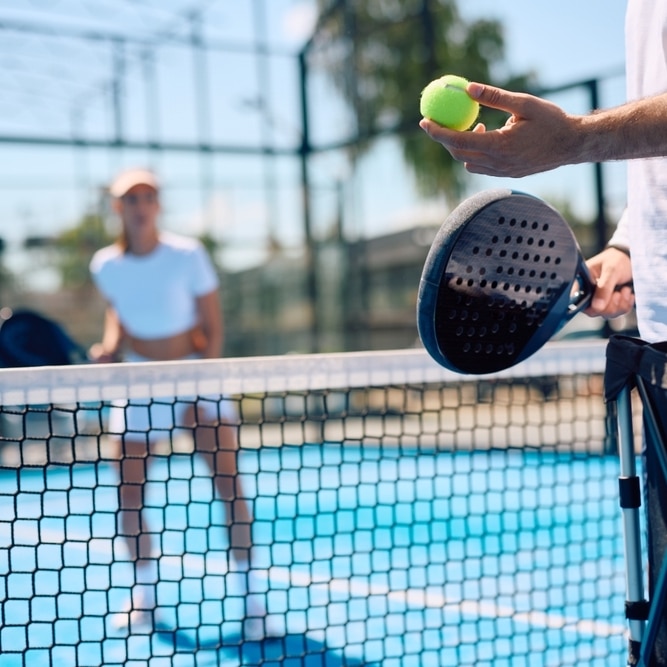 Padel Tennis : Entraînement et Match sur Court Bleu Main tenant une raquette et une balle de padel sur un court bleu avec une joueuse de tennis en arrière-plan.